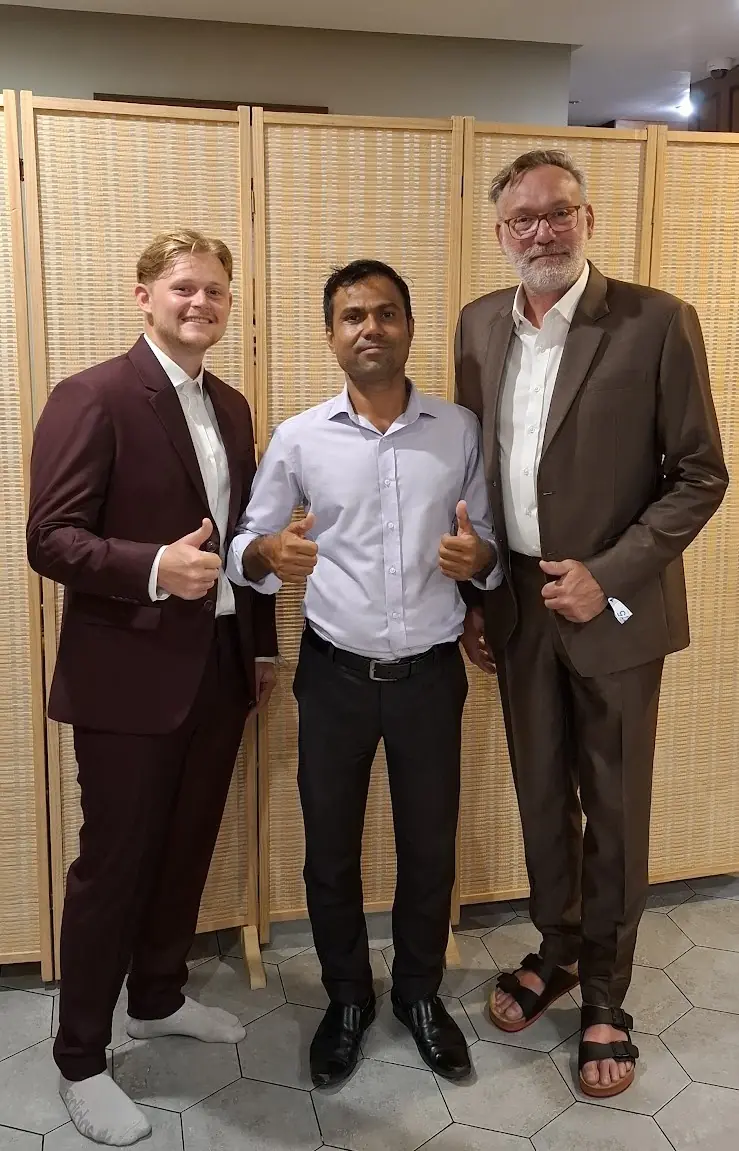 Two finished suits — burgundy and brown — with the tailor who made them, photographed at the customers' home.