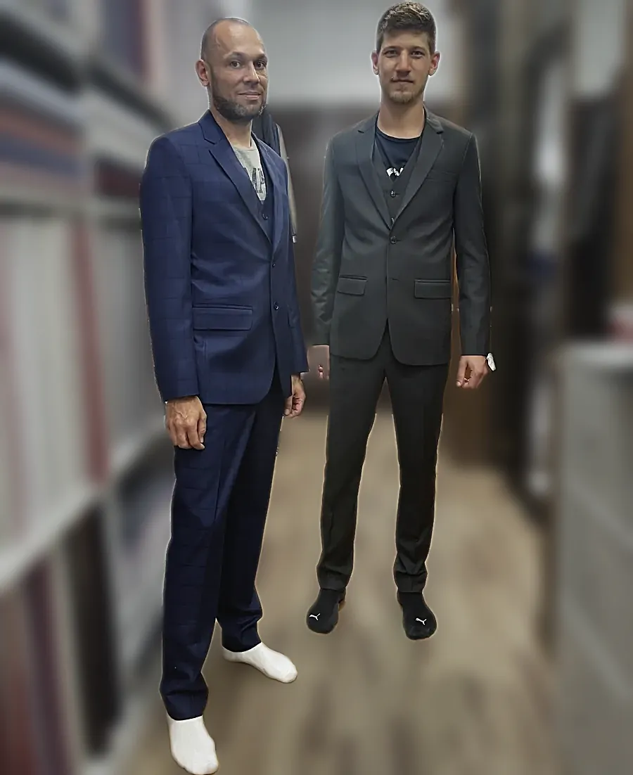 Two three-piece suits at the fitting — a navy windowpane and a dark grey, both with their waistcoats on.
