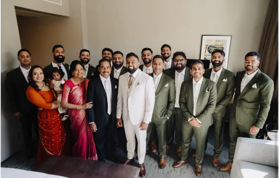 The whole wedding party with family — the groom in cream, the groomsmen in olive, in the hotel suite.
