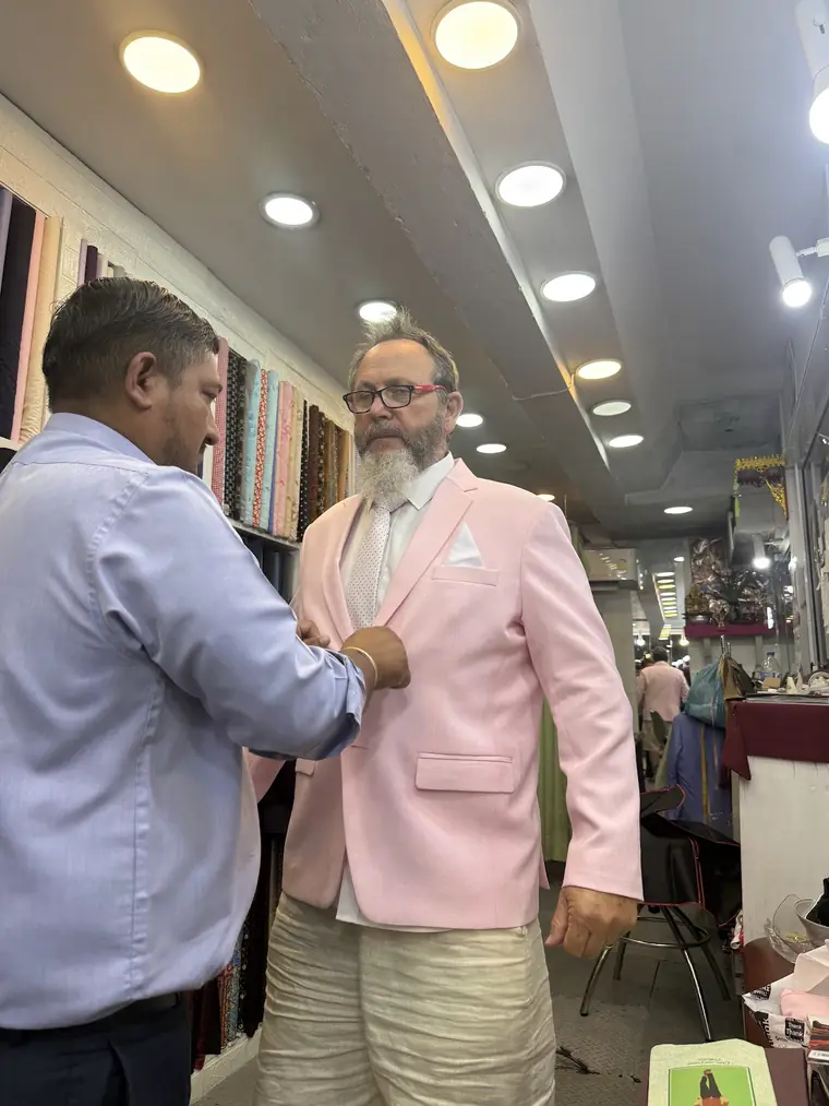 A tailor fastening the front of the pale pink jacket the reviewer asked for, worn with cream trousers and a white pocket square, in front of the shirting racks.