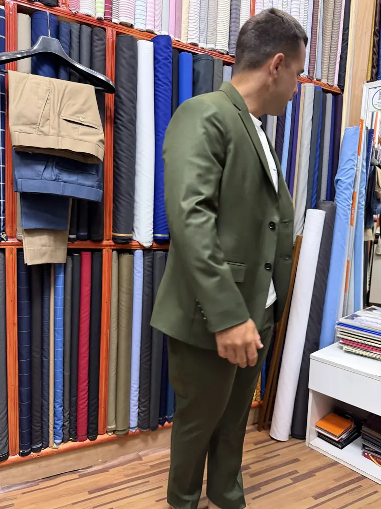 The reviewer in profile in the plain olive-green two-piece they had made, against floor-to-ceiling racks of suiting and shirting.