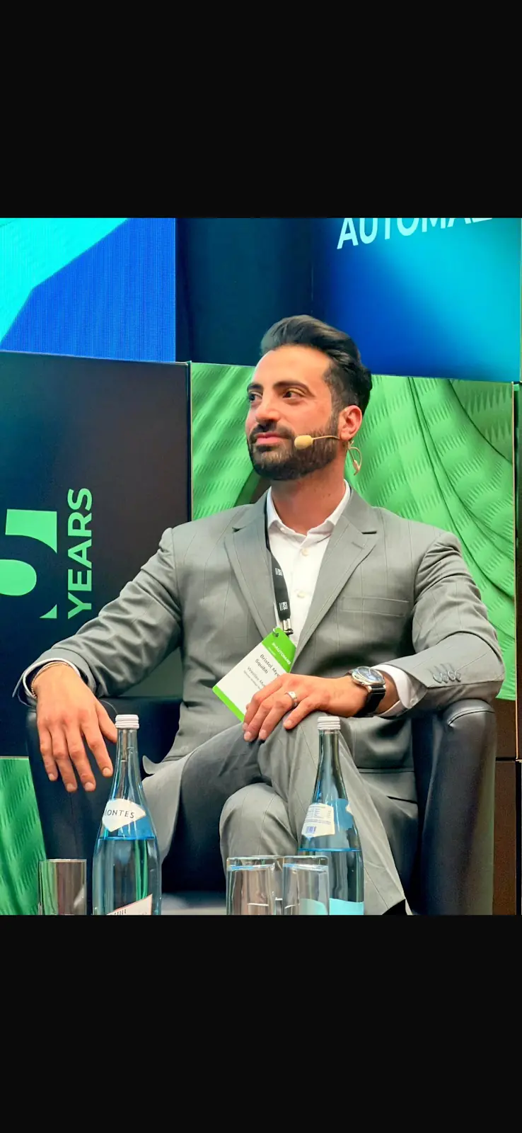 The reviewer wearing their grey suit on a conference panel, open-collar white shirt and lanyard, under the stage lights.
