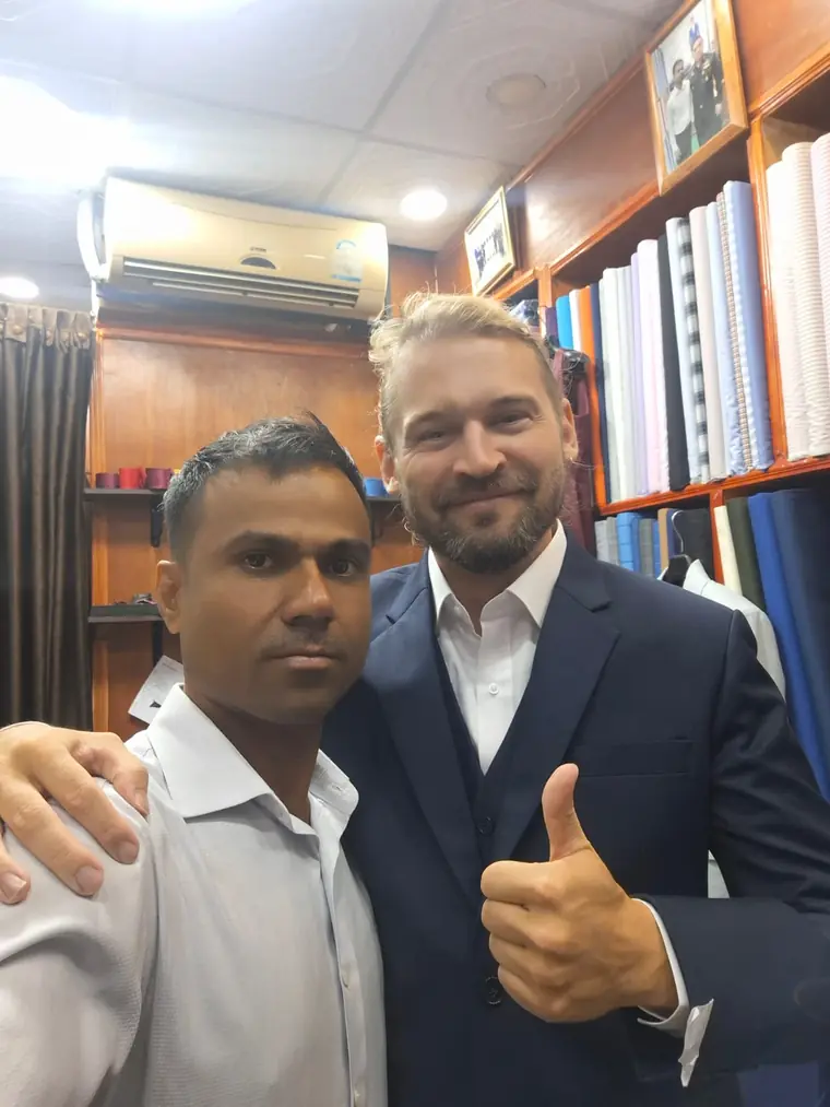 The reviewer in a navy suit and white shirt, thumbs up in a selfie with one of the tailors, shirting bolts shelved behind them.