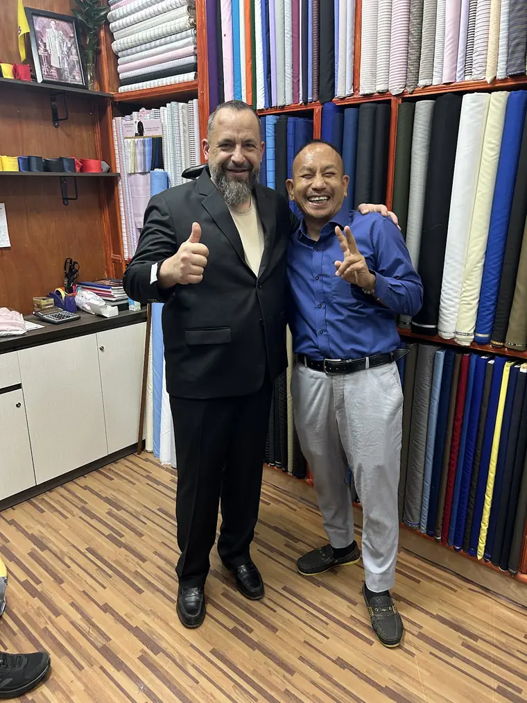 The reviewer in the black suit they had made, thumbs up beside one of the tailors, the walls of shirting and suiting bolts behind them.