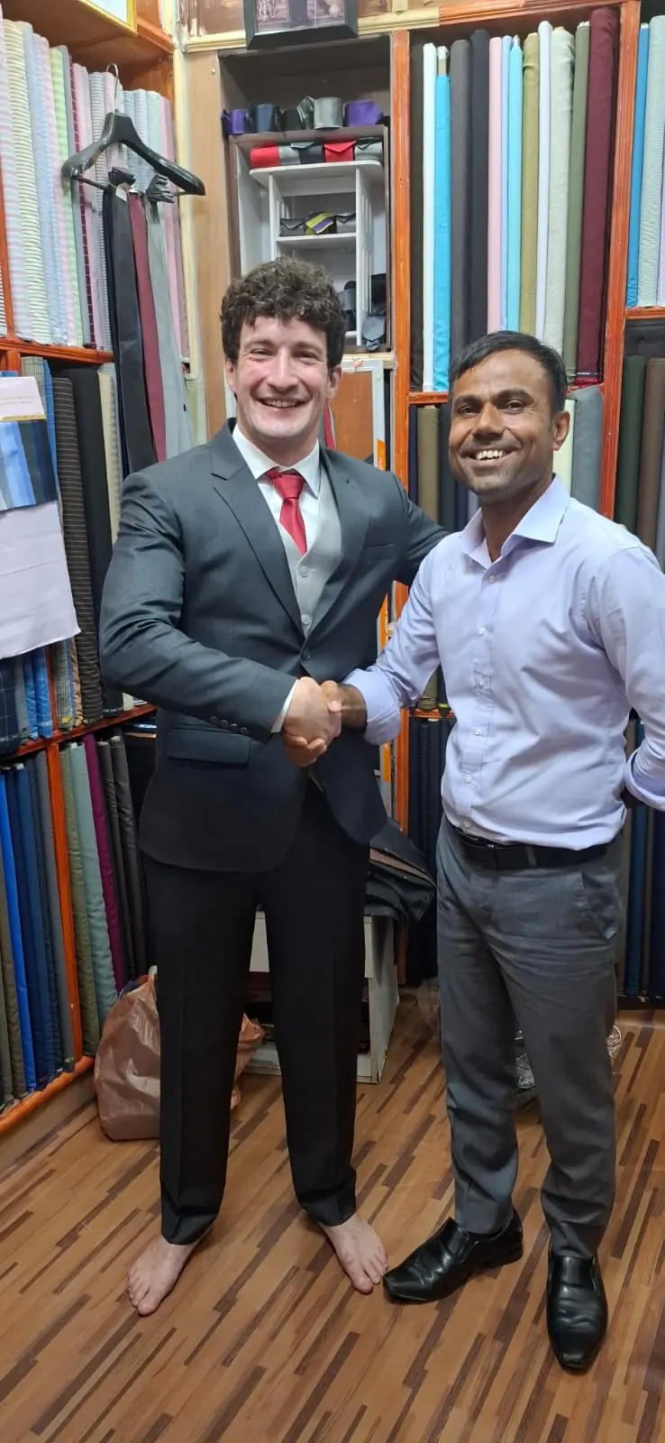 The reviewer shaking hands with one of the tailors, wearing the charcoal three-piece and red tie they had made for a wedding, standing barefoot while the trousers were finished.