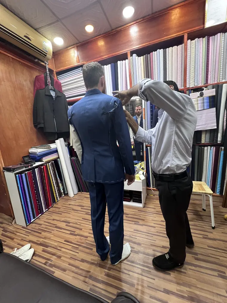 A basted fitting in progress: the reviewer in a blue jacket still held together with white tacking stitches, while a tailor works on the sleeve head.