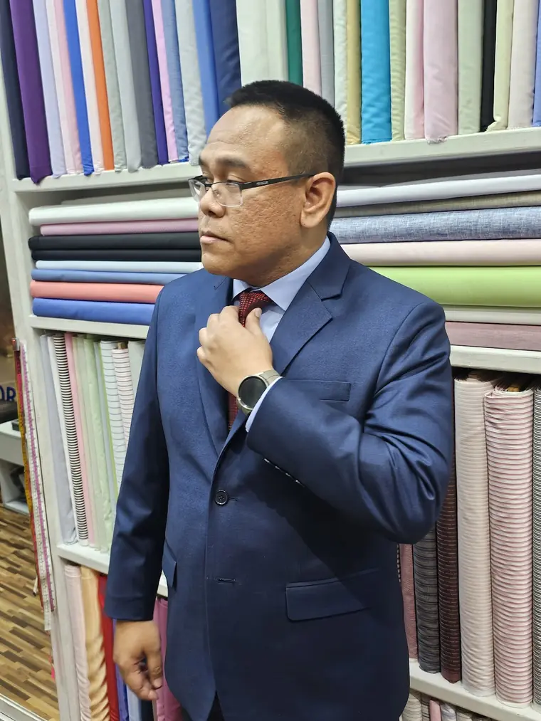The reviewer straightening a burgundy tie in the blue suit they had made, against shelves of pastel shirting.