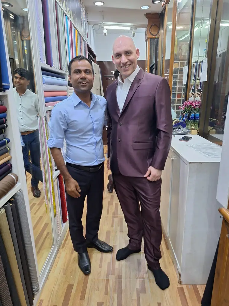 The reviewer in the plum sharkskin suit they had made, standing in socks beside one of the tailors in the fitting aisle.