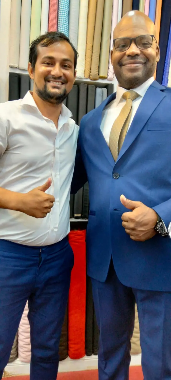 The reviewer in a royal-blue suit and gold tie, thumbs up beside one of the tailors, the cloth shelves behind them.