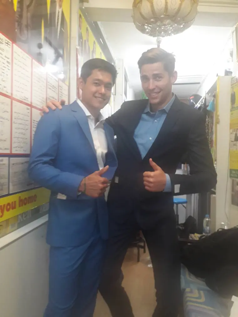 Two finished suits, one bright blue and one black, thumbs up in front of the wall of handwritten notes customers have left.