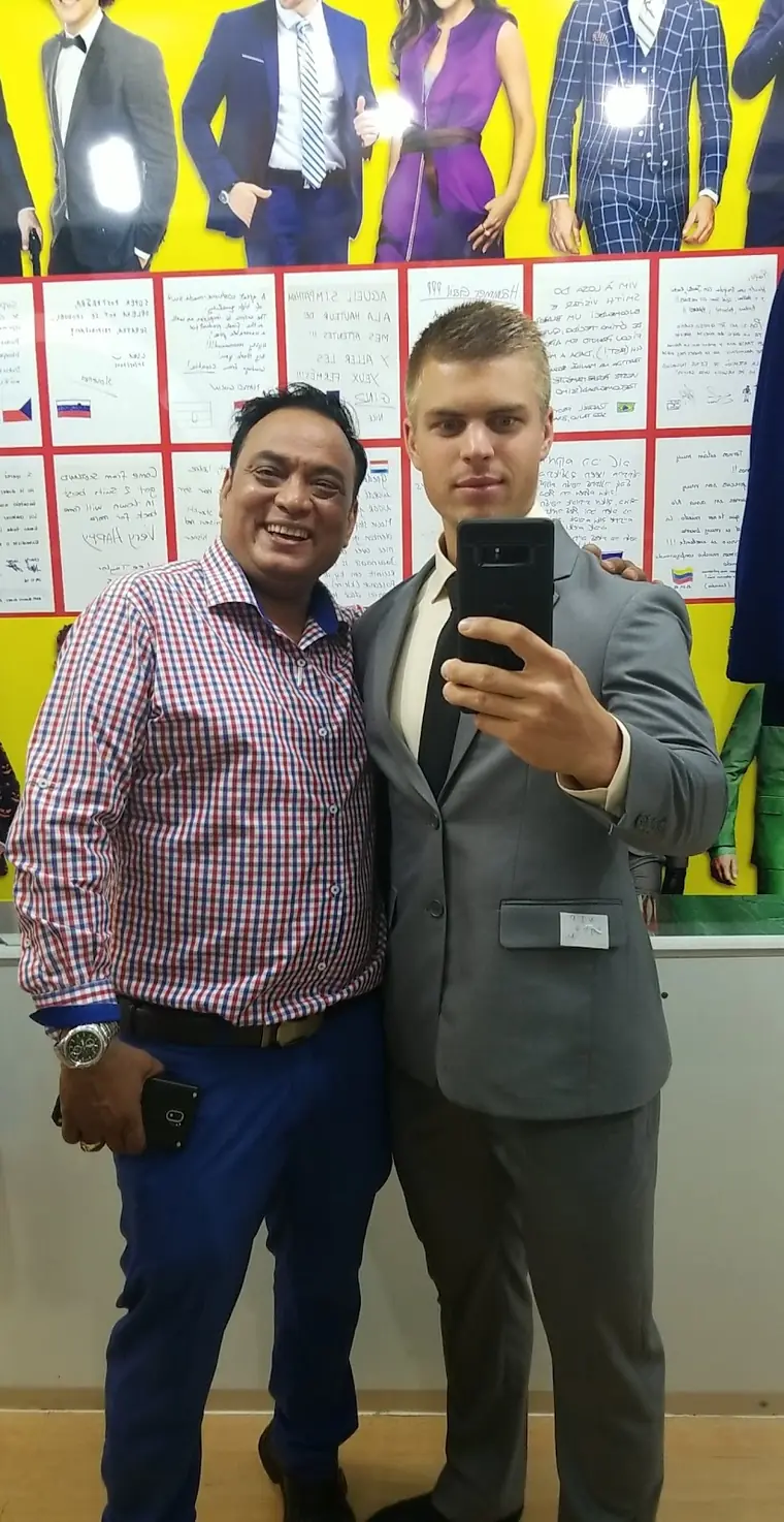 A mirror selfie in a grey suit with the maker’s tag still on the pocket, taken with one of the tailors in front of the wall of customer notes.