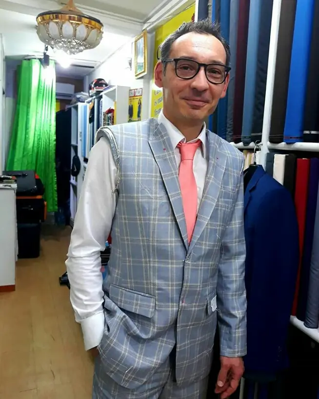 The reviewer in a pale blue windowpane-check suit and coral tie, the maker’s tag still on the pocket, standing among the racks of cloth.