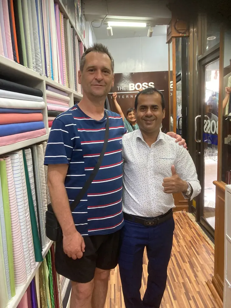 The reviewer photographed with one of the tailors beside the wall of shirting bolts, the shop mirror and signage behind them.