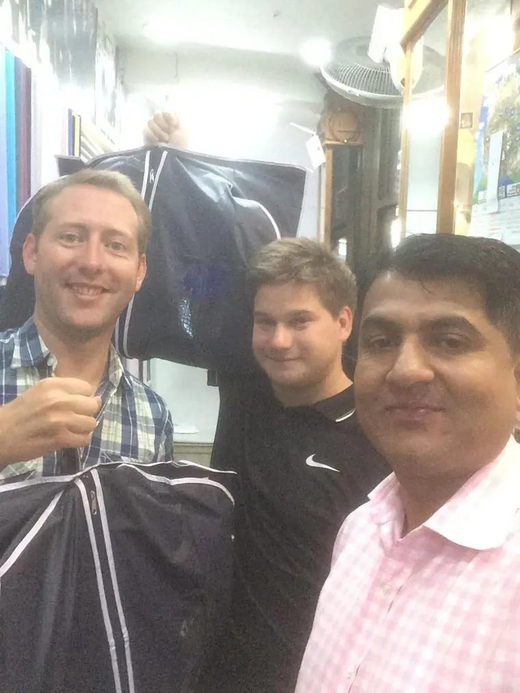 The reviewer holding up a finished garment in its carrier, photographed with a companion and one of the tailors.