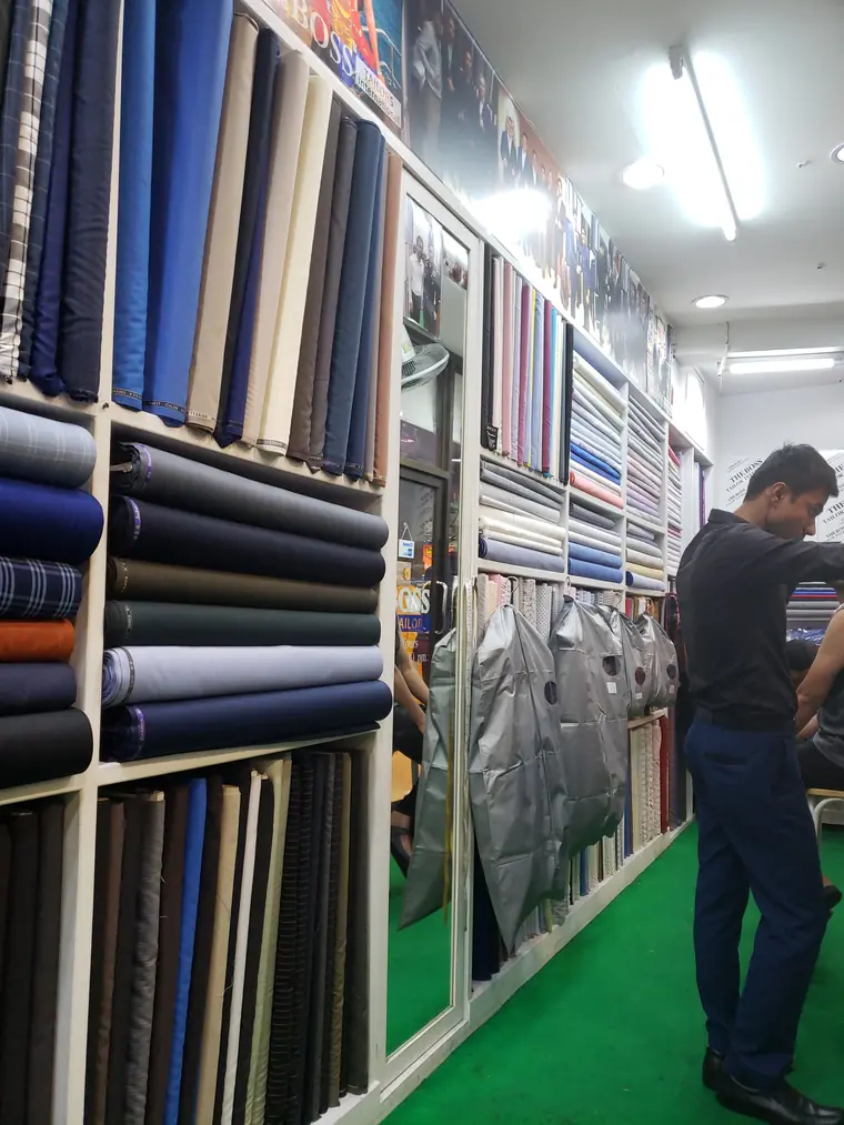 The cloth wall: shelved bolts of suiting and shirting from floor to ceiling, with finished garments hanging in their carriers.